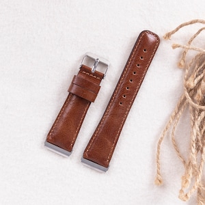 May include: Two brown leather watch straps with silver buckles. The straps have stitching along the edges and are laid out on a white surface.