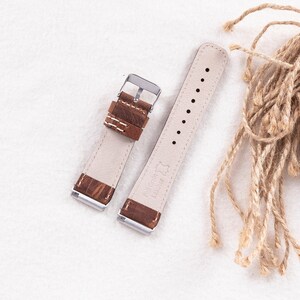 Puede incluir: Dos correas de reloj de cuero marr&oacute;n con hebillas plateadas. Las correas tienen costuras blancas y est&aacute;n dispuestas sobre una superficie blanca.