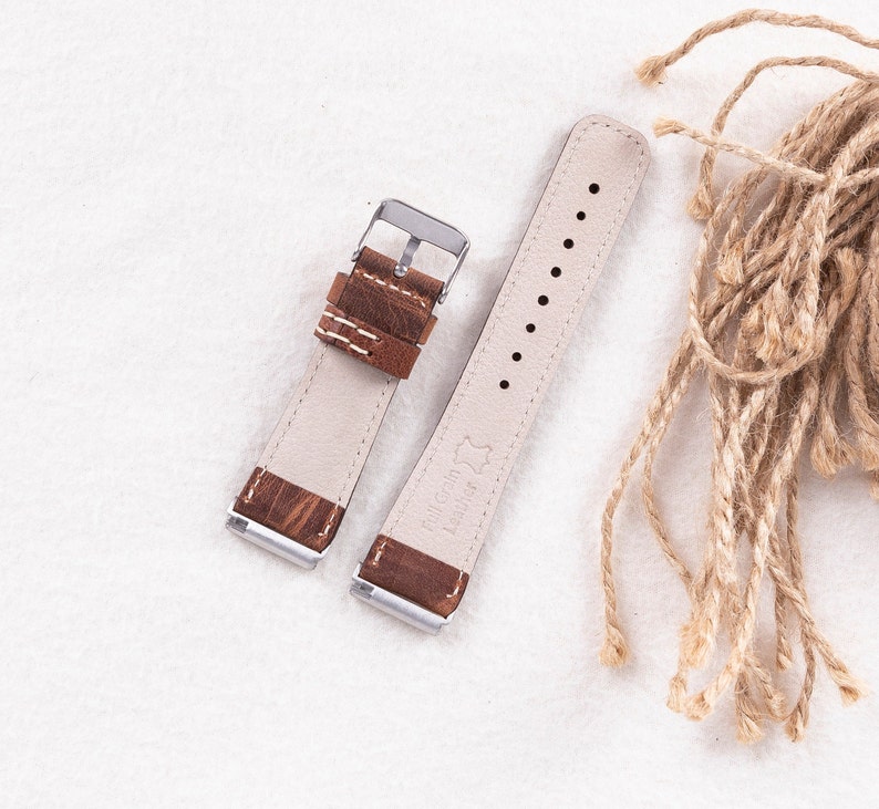 Puede incluir: Dos correas de reloj de cuero marr&oacute;n con hebillas plateadas. Las correas tienen costuras blancas y est&aacute;n dispuestas sobre una superficie blanca.