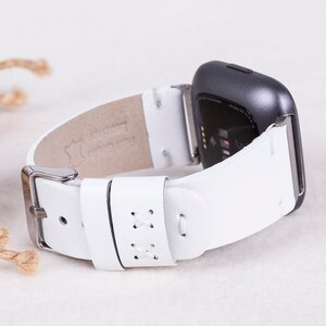 以下が含まれることがあります： Fitbitスマートウォッチ用に設計された、ステッチのディテールが施された白い革製の腕時計バンド。バンドには銀色のバックルが付いています。