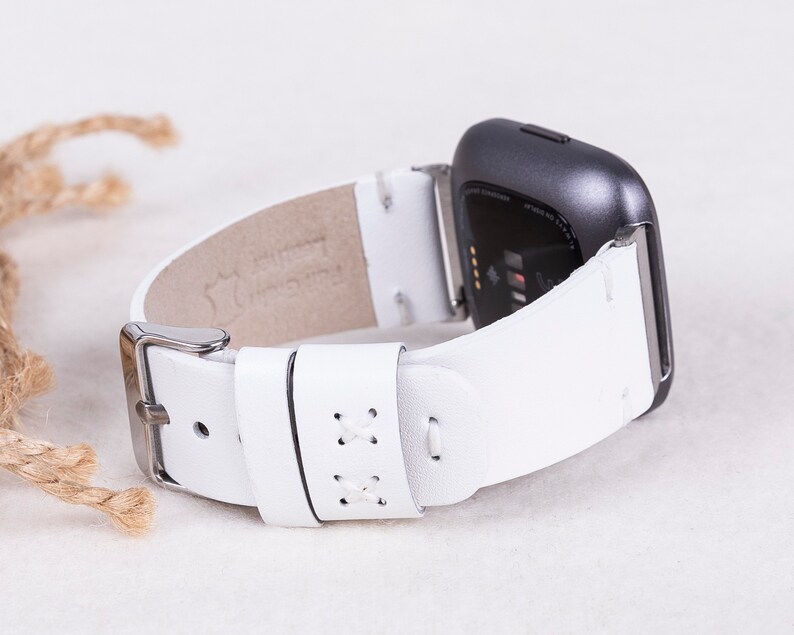 以下が含まれることがあります： Fitbitスマートウォッチ用に設計された、ステッチのディテールが施された白い革製の腕時計バンド。バンドには銀色のバックルが付いています。