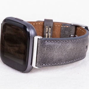 May include: A gray leather watch band with a silver buckle. The band is designed for a smartwatch with a square face.