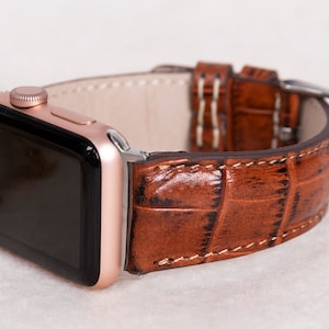 May include: A rose gold smartwatch with a brown leather strap. The strap has a crocodile skin texture and is secured with a silver buckle. The watch face is black.