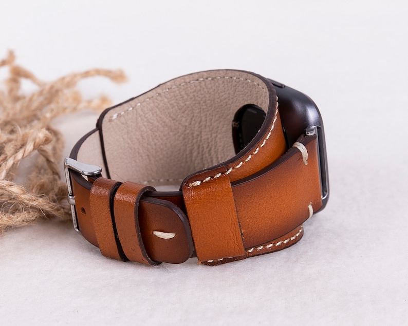 Puede incluir: Una correa de reloj de cuero marr&oacute;n con costuras blancas y una hebilla plateada. La correa est&aacute; dise&ntilde;ada para adaptarse a un reloj inteligente.