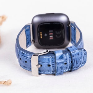 Puede incluir: Una correa de reloj de cuero azul claro con una hebilla plateada. La correa est&aacute; dise&ntilde;ada para un reloj inteligente Fitbit.