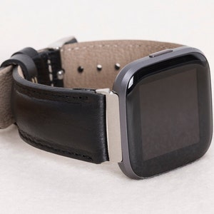 May include: A black leather watch band with a silver buckle is attached to a gray smartwatch. The watch band is made of smooth, black leather and has a light gray underside. The watch face is black and rectangular.