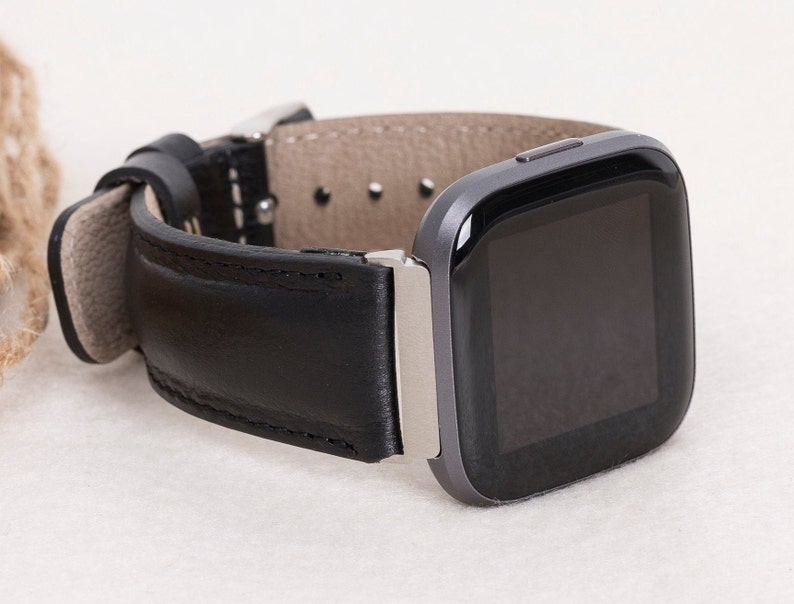 May include: A black leather watch band with a silver buckle is attached to a gray smartwatch. The watch band is made of smooth, black leather and has a light gray underside. The watch face is black and rectangular.