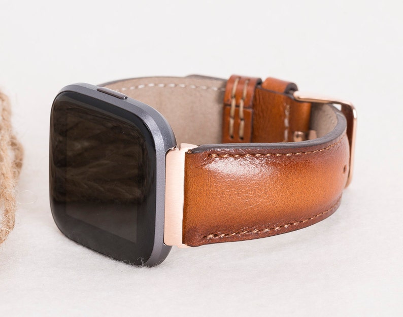 Op de afbeelding: Een bruin leren horlogebandje met een ros&eacute;gouden gesp en stiksels. Het bandje is bevestigd aan een vierkant smartwatch met een zwart scherm.