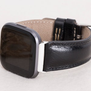 May include: A black leather watch band with a silver buckle is attached to a gray smartwatch. The watch band has white stitching and a light brown interior. The watch face is black and the watch is on a light background.