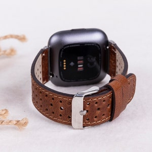 May include: Brown leather watch band with a perforated design and a silver buckle. The band is compatible with a Fitbit smartwatch.