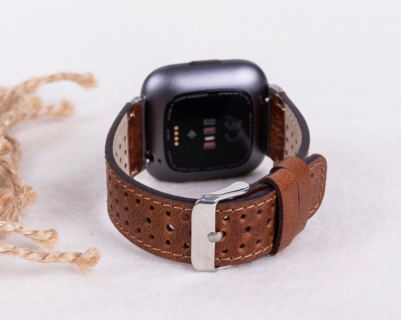 May include: Brown leather watch band with a perforated design and a silver buckle. The band is compatible with a Fitbit smartwatch.