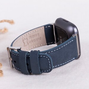 May include: A dark blue leather watch band with white stitching. The band is attached to a silver buckle and has the words "Full Grain Leather" printed on the underside.