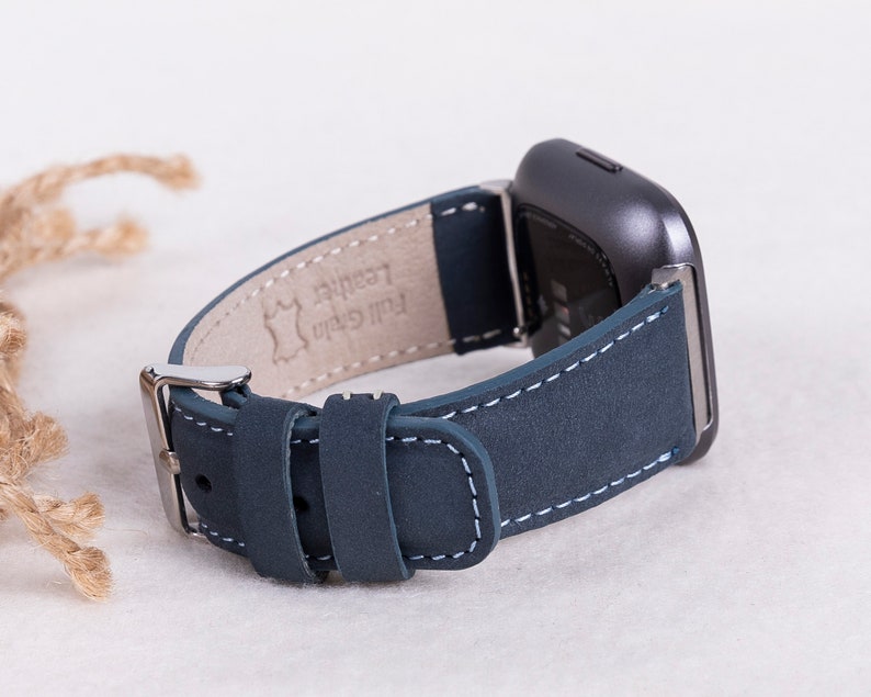 May include: A dark blue leather watch band with white stitching. The band is attached to a silver buckle and has the words "Full Grain Leather" printed on the underside.