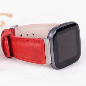 Puede incluir: Una correa de reloj de cuero rojo con una hebilla plateada, unida a un reloj inteligente negro con una cara cuadrada.