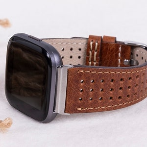 May include: A brown leather watch band with white stitching and a perforated design. The band is compatible with a smartwatch.