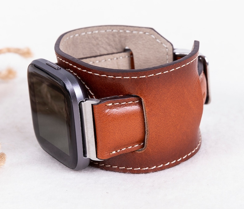 K&ouml;nnte beinhalten: Ein braunes Lederarmband mit wei&szlig;en N&auml;hten. Das Armband ist f&uuml;r eine quadratische Smartwatch konzipiert.
