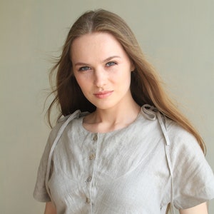 Puede incluir: Una camisa de lino gris claro con botones, mangas cortas y detalles de lazo en los hombros. La camisa tiene un cuello redondo y se abotona por delante. La modelo lleva la camisa.