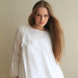 Può includere: Una tunica bianca in lino con scollo arricciato e maniche lunghe. La tunica ha una vestibilità rilassata e una forma leggermente a trapezio. L'orlo è rifinito con un sottile bordo grigio.