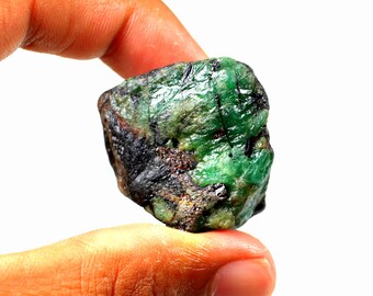 May include: A rough, green emerald gemstone with black and brown inclusions. The emerald is held in a hand.