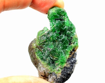 May include: A rough, green emerald crystal with black inclusions. The crystal is translucent and has a glassy luster.