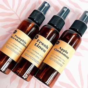 May include: Three 2-ounce amber glass bottles with black spray tops and labels. The labels are brown with gold accents and feature the names of the scents: Pumpkin Marshmallow, Fresh Linen, and Apple Cinnamon. Each label also includes the text "Daisy Garden Gifts - Etsy" and "Handmade in Ohio".
