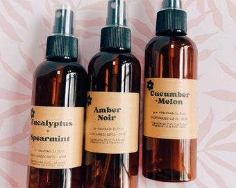 May include: Three amber glass spray bottles with black spray tops and labels. The labels are brown with white text and a flower graphic. The labels read "Eucalyptus + Spearmint", "Amber Noir", and "Cucumber + Melon".