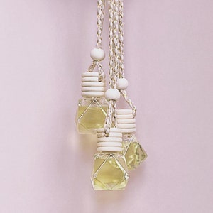K&ouml;nnte beinhalten: Vier kleine, klare, geometrisch geformte Glasflaschen, gef&uuml;llt mit gelber Fl&uuml;ssigkeit, h&auml;ngen an geflochtenen wei&szlig;en und goldenen Kordeln. Jede Flasche hat einen Holzverschluss und eine Holzperle. Der Hintergrund ist ein hellrosa Farbton.