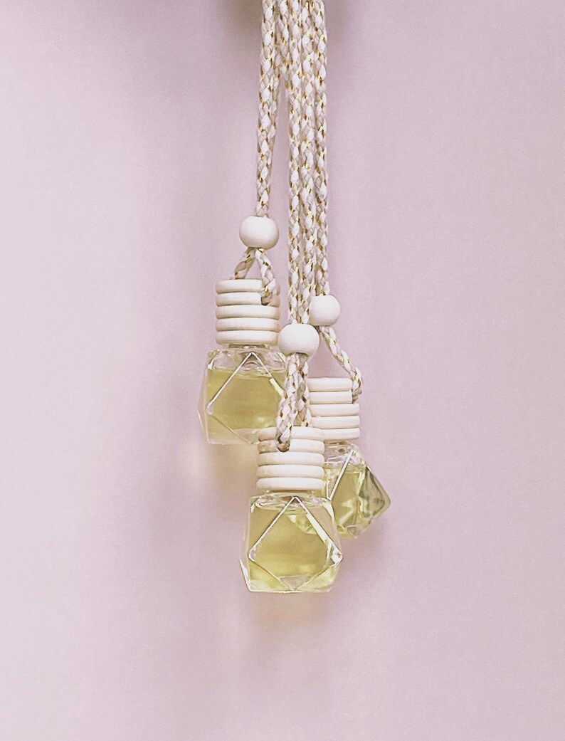 K&ouml;nnte beinhalten: Vier kleine, klare, geometrisch geformte Glasflaschen, gef&uuml;llt mit gelber Fl&uuml;ssigkeit, h&auml;ngen an geflochtenen wei&szlig;en und goldenen Kordeln. Jede Flasche hat einen Holzverschluss und eine Holzperle. Der Hintergrund ist ein hellrosa Farbton.