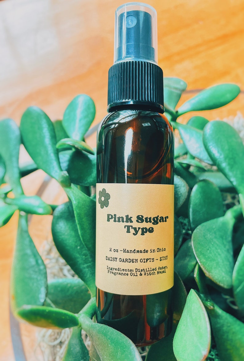 May include: A brown glass bottle with a black spray top and a label that reads "Pink Sugar Type" with the text "2 oz - Handmade in Ohio Daisy Garden Gifts - Etsy Ingredients: Distilled Water, Fragrance Oil & Witch Hazel".