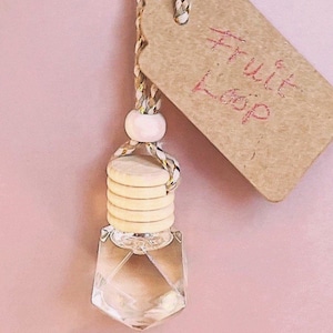 K&ouml;nnte beinhalten: Eine klare, geometrisch geformte Glasflasche mit einem Holzverschluss und einem Etikett mit der Aufschrift "Fruit Loop". Die Flasche wird von einer geflochtenen Schnur mit einer Holzperle gehalten. Der Hintergrund ist einfarbig rosa.