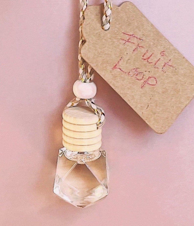K&ouml;nnte beinhalten: Eine klare, geometrisch geformte Glasflasche mit einem Holzverschluss und einem Etikett mit der Aufschrift "Fruit Loop". Die Flasche wird von einer geflochtenen Schnur mit einer Holzperle gehalten. Der Hintergrund ist einfarbig rosa.
