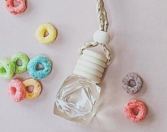 May include: A clear, square-shaped car air freshener with a white wooden top and a braided rope for hanging. Colorful, round cereal pieces in pink, yellow, green, blue, and purple are scattered around the freshener on a light pink surface.