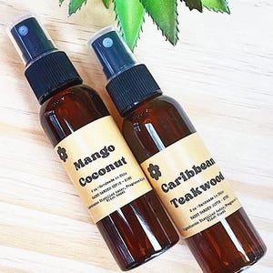 May include: Two brown glass spray bottles with brown labels. The bottle on the left is labeled "Mango Coconut" and the bottle on the right is labeled "Caribbean Teakwood". Both bottles have a small flower icon on the label.