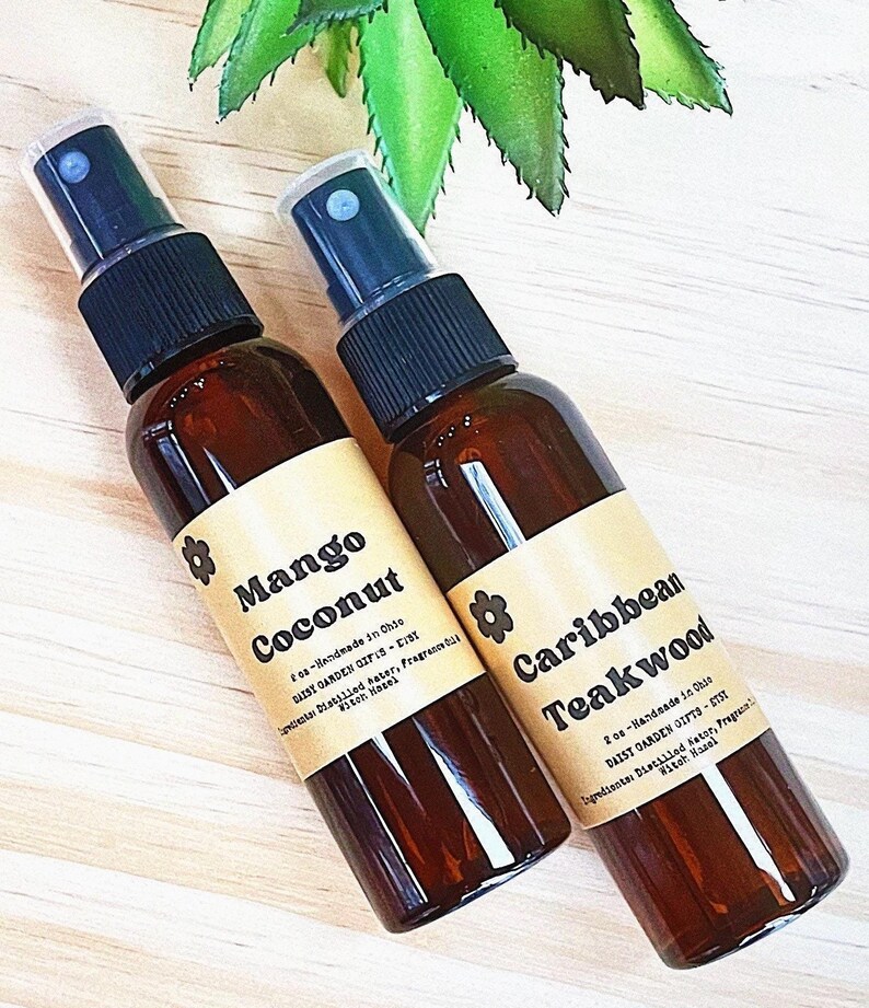May include: Two brown glass spray bottles with brown labels. The bottle on the left is labeled "Mango Coconut" and the bottle on the right is labeled "Caribbean Teakwood". Both bottles have a small flower icon on the label.