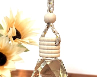 May include: A clear, diamond-shaped glass bottle hanging from a braided white and gold cord. The bottle features a light-colored wooden cap with horizontal grooves and a single wooden bead above it, suspended by a loop of the same cord.
