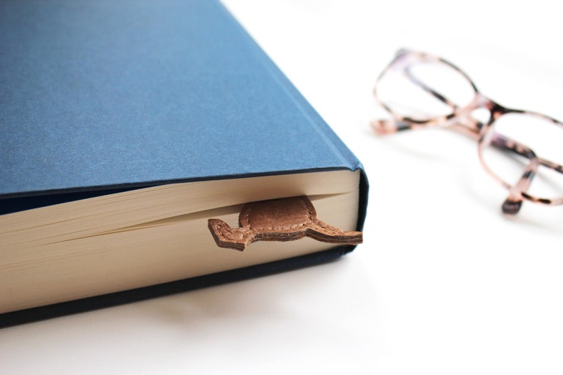 K&ouml;nnte beinhalten: Ein blaues Hardcover-Buch mit einem braunen Leder-Lesezeichen in Stiefelform. Das Lesezeichen befindet sich zwischen den Seiten. Eine Brille mit Schildpattmuster liegt im Hintergrund.
