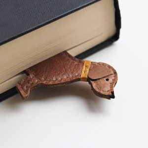 The top of the leather dachshund bookmark peaks out of the book, the bookmark is a tan brown colour and had the face in side profile of a sausage dog with ears, eyes and leather collar