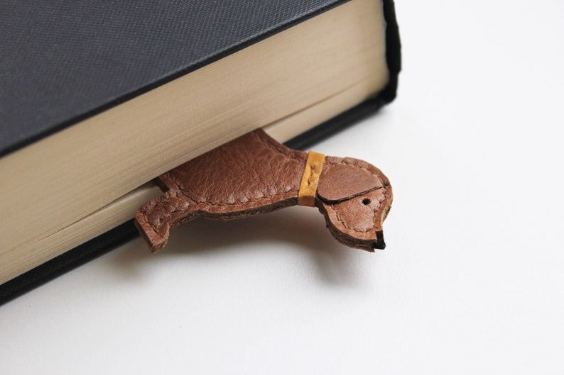 The top of the leather dachshund bookmark peaks out of the book, the bookmark is a tan brown colour and had the face in side profile of a sausage dog with ears, eyes and leather collar