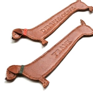K&ouml;nnte beinhalten: Zwei braune Leder-Dackel-Schl&uuml;sselanh&auml;nger. Einer hat den gepr&auml;gten Namen "DENNY & OLIVE", der andere "FRANK". Jeder hat ein farbiges Halsband: rot und gr&uuml;n.