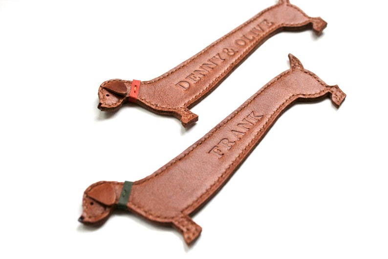 K&ouml;nnte beinhalten: Zwei braune Leder-Dackel-Schl&uuml;sselanh&auml;nger. Einer hat den gepr&auml;gten Namen "DENNY & OLIVE", der andere "FRANK". Jeder hat ein farbiges Halsband: rot und gr&uuml;n.