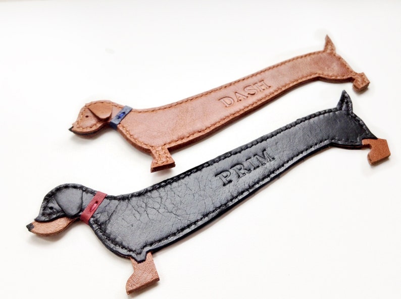 K&ouml;nnte beinhalten: Zwei Lesezeichen aus Leder in Dackelform. Eines ist hellbraun mit dem Pr&auml;gedruck "DASH", das andere ist schwarz mit dem Pr&auml;gedruck "PRIM". Beide haben kontrastierende Kragen.