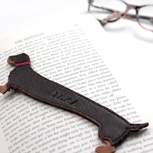 May include: A brown leather dachshund bookmark with the name "ILKA" embossed on it. The bookmark is lying on a book with a pair of glasses on top.