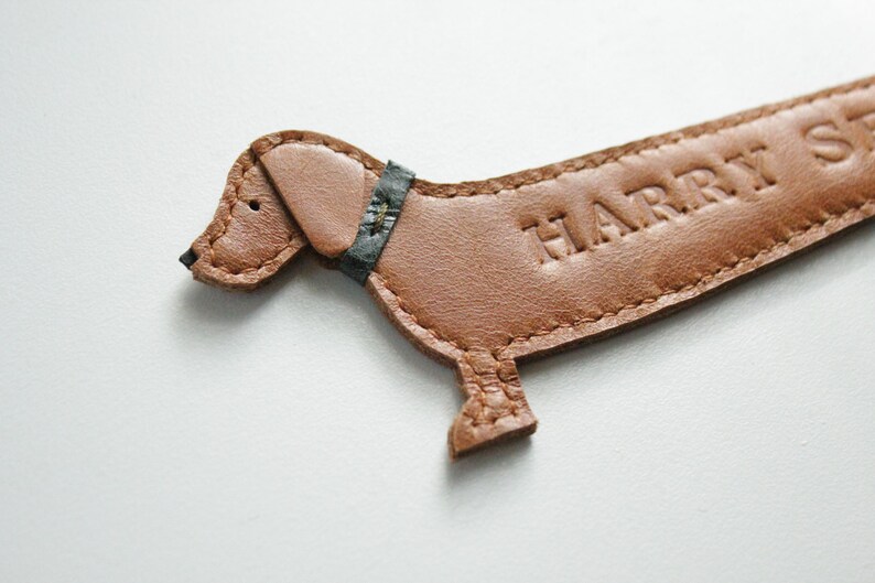 K&ouml;nnte beinhalten: Ein brauner Lederanh&auml;nger in Dackelform mit dem gepr&auml;gten Namen "HARRY ST" auf der Seite. Der Hund hat eine schwarze Nase, einen dunkelblauen Kragen und ist an den R&auml;ndern gen&auml;ht.