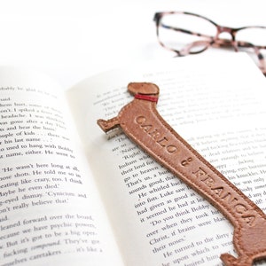 A mid brown dachshund shaped leather bookmark rests on a book, as dachshunds are long dogs the bookmark is exaggeratedly stretched and embossed with custom wording and is saddle stitched together featuring ears and collar detailing