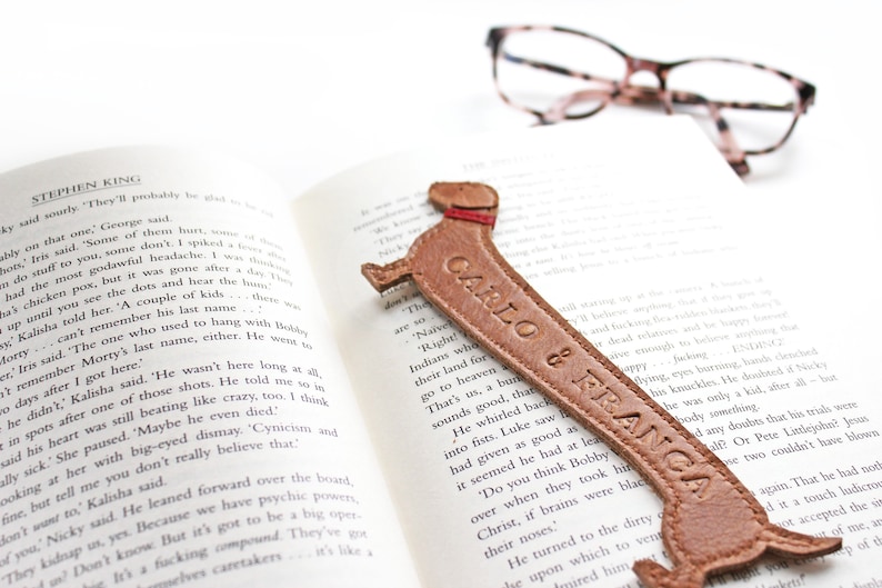 A mid brown dachshund shaped leather bookmark rests on a book, as dachshunds are long dogs the bookmark is exaggeratedly stretched and embossed with custom wording and is saddle stitched together featuring ears and collar detailing
