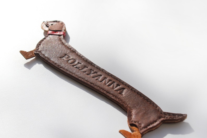 A two tone dark and mid brown sausage dog shaped leather bookmark which is sewn together and has text embossed along its body, this text can be customised along with the colour of this bookmark