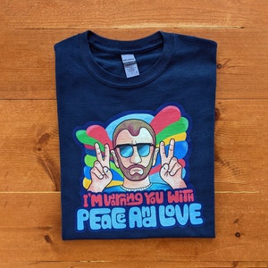 Puede incluir: Una camiseta azul marino con un gráfico colorido de un hombre con gafas de sol y barba haciendo el signo de la paz. El texto "I'm warming you with peace and love" está impreso debajo del gráfico.