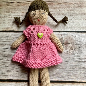May include: Hand-knitted doll with brown hair in pigtails and a pink dress. The doll has a tan face and limbs, with a green heart-shaped button on the dress. The doll is made of yarn and sits on a wooden surface.