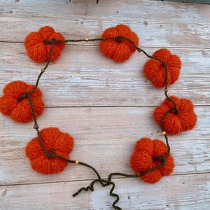 May include: A circular garland of seven knitted orange pumpkins, ideal for autumn decorations. The pumpkins are linked by a dark green string with small wooden beads. The garland is a charming, handmade piece.