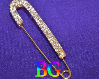 May include: A gold-colored safety pin with a row of clear rhinestones along the curved top and a portion of the pin. The pin is set against a purple background. The letters "BG" are visible in the image.